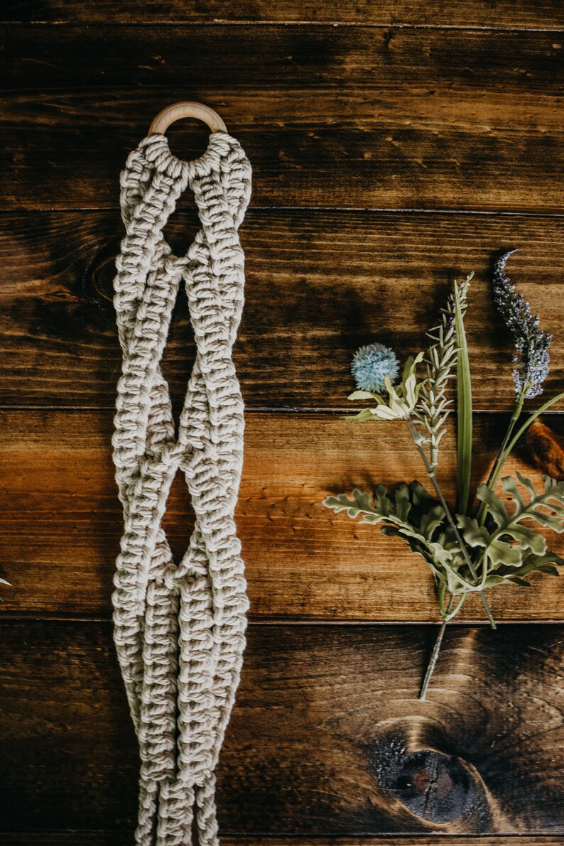 May include: A beige macrame plant hanger with a wooden ring at the top. The hanger is hanging against a dark brown wooden background.