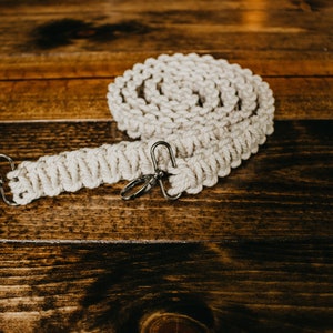 May include: A white cotton macrame camera strap with silver metal clasps. The strap is coiled on a wooden surface.