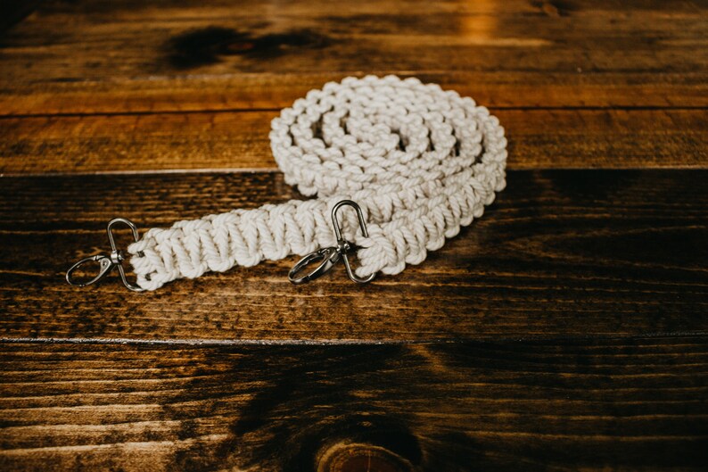May include: A white cotton macrame camera strap with silver metal clasps. The strap is coiled on a wooden surface.