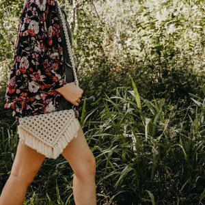May include: A woman wearing a floral kimono and a white macrame bag with fringe. The bag is worn over her shoulder and is a triangular shape.