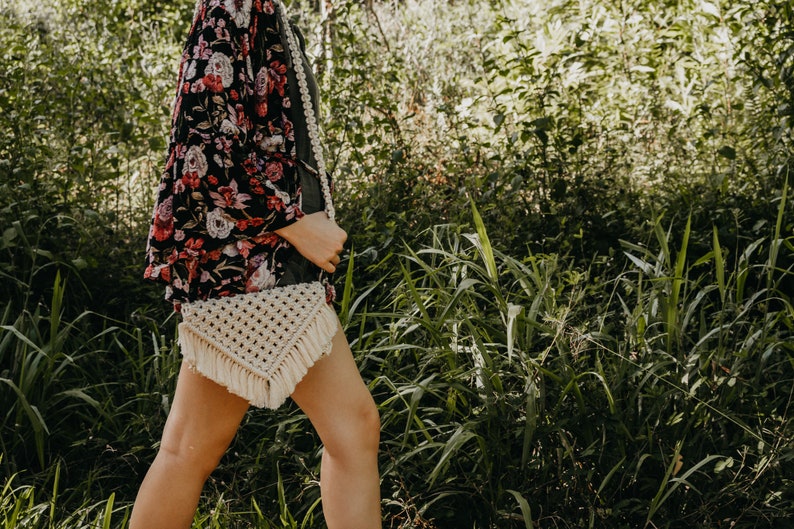 May include: A woman wearing a floral kimono and a white macrame bag with fringe. The bag is worn over her shoulder and is a triangular shape.