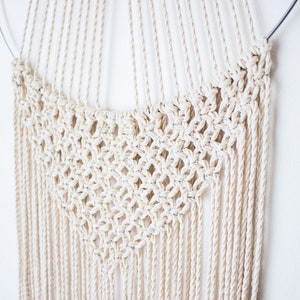 Op de afbeelding: Cr&egrave;mekleurige macram&eacute; wandhanger. Het ontwerp heeft een cirkelvormige bovenkant met een geweven ruitpatroon in het midden en lange, verticale franjes. Het stuk is gemaakt van natuurlijk gekleurd katoenen touw en hangt tegen een witte muur.