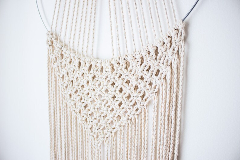 Op de afbeelding: Cr&egrave;mekleurige macram&eacute; wandhanger. Het ontwerp heeft een cirkelvormige bovenkant met een geweven ruitpatroon in het midden en lange, verticale franjes. Het stuk is gemaakt van natuurlijk gekleurd katoenen touw en hangt tegen een witte muur.