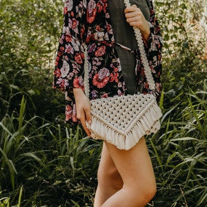 May include: A white macrame crossbody bag with fringe detailing. The bag is worn by a person wearing a floral print kimono and a dark green dress.