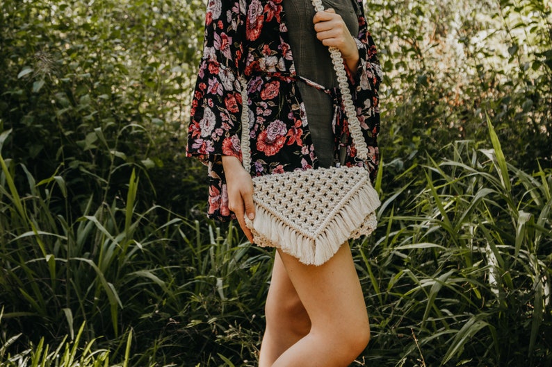 May include: A white macrame crossbody bag with fringe detailing. The bag is worn by a person wearing a floral print kimono and a dark green dress.