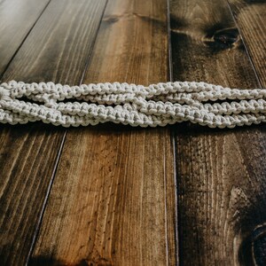 May include: A white macrame plant hanger with a wooden ring. The hanger is lying on a dark brown wooden surface.