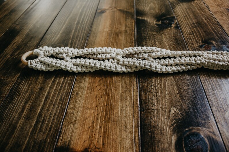 May include: A white macrame plant hanger with a wooden ring. The hanger is lying on a dark brown wooden surface.
