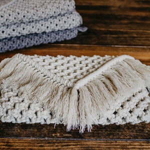 May include: A beige crocheted throw blanket with fringe on all sides. The blanket is folded in half and lying on a wooden surface.