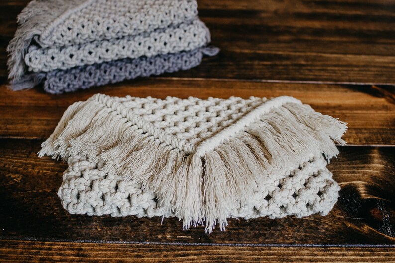 May include: A beige crocheted throw blanket with fringe on all sides. The blanket is folded in half and lying on a wooden surface.