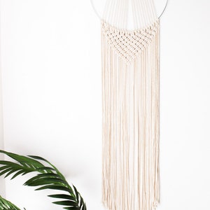 Op de afbeelding: Een cr&egrave;mekleurige macram&eacute; wandhanger. Het ontwerp heeft een geweven bovenste gedeelte met een geometrisch patroon, opgehangen aan een zilveren metalen ring. Lange, gefranjerde strengen van hetzelfde cr&egrave;mekleurige materiaal vallen naar beneden.