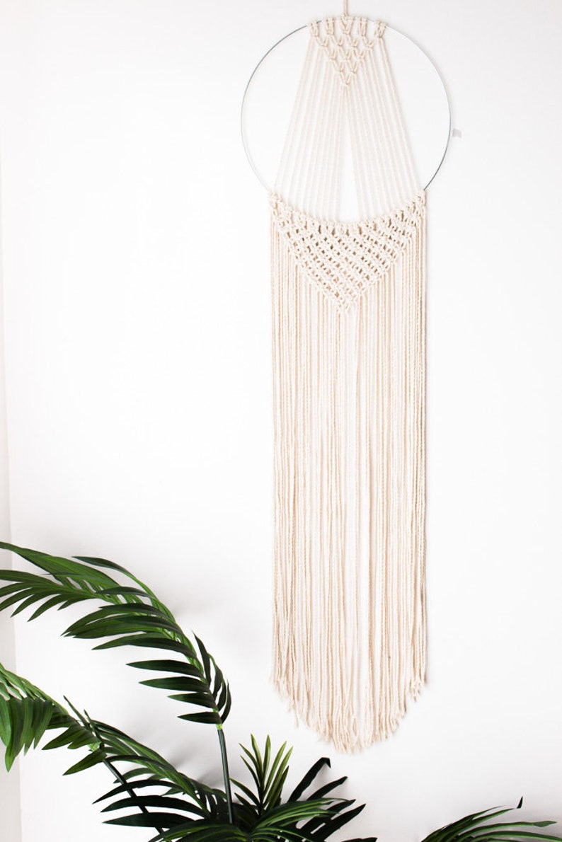 Op de afbeelding: Een cr&egrave;mekleurige macram&eacute; wandhanger. Het ontwerp heeft een geweven bovenste gedeelte met een geometrisch patroon, opgehangen aan een zilveren metalen ring. Lange, gefranjerde strengen van hetzelfde cr&egrave;mekleurige materiaal vallen naar beneden.