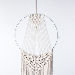 Op de afbeelding: Een macram&eacute; wandhanger met een cirkelvormig metalen frame en ingewikkelde geweven patronen. De cr&egrave;mekleurige katoenen koorden zijn in een geometrisch ontwerp geknoopt, waardoor een textuurrijke, bohemian-stijl woondecoratie ontstaat.