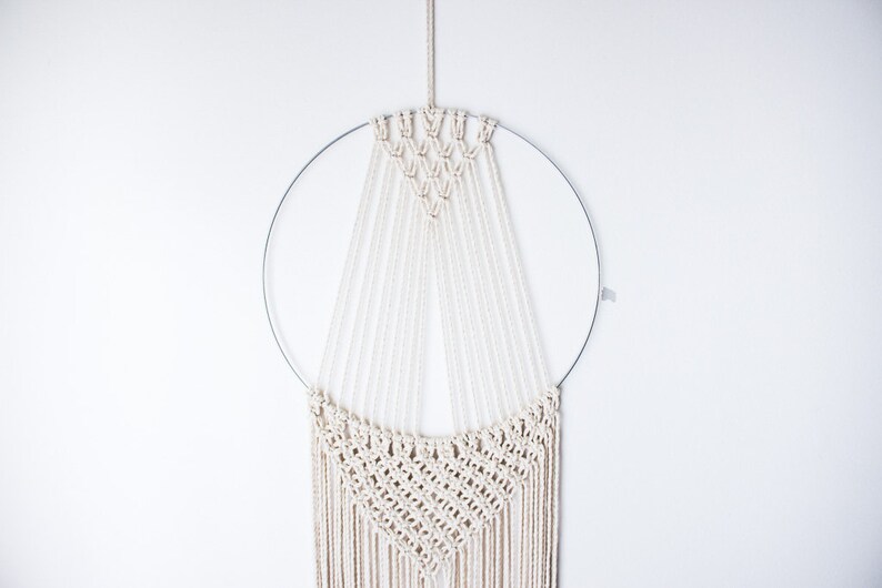 Op de afbeelding: Een macram&eacute; wandhanger met een cirkelvormig metalen frame en ingewikkelde geweven patronen. De cr&egrave;mekleurige katoenen koorden zijn in een geometrisch ontwerp geknoopt, waardoor een textuurrijke, bohemian-stijl woondecoratie ontstaat.