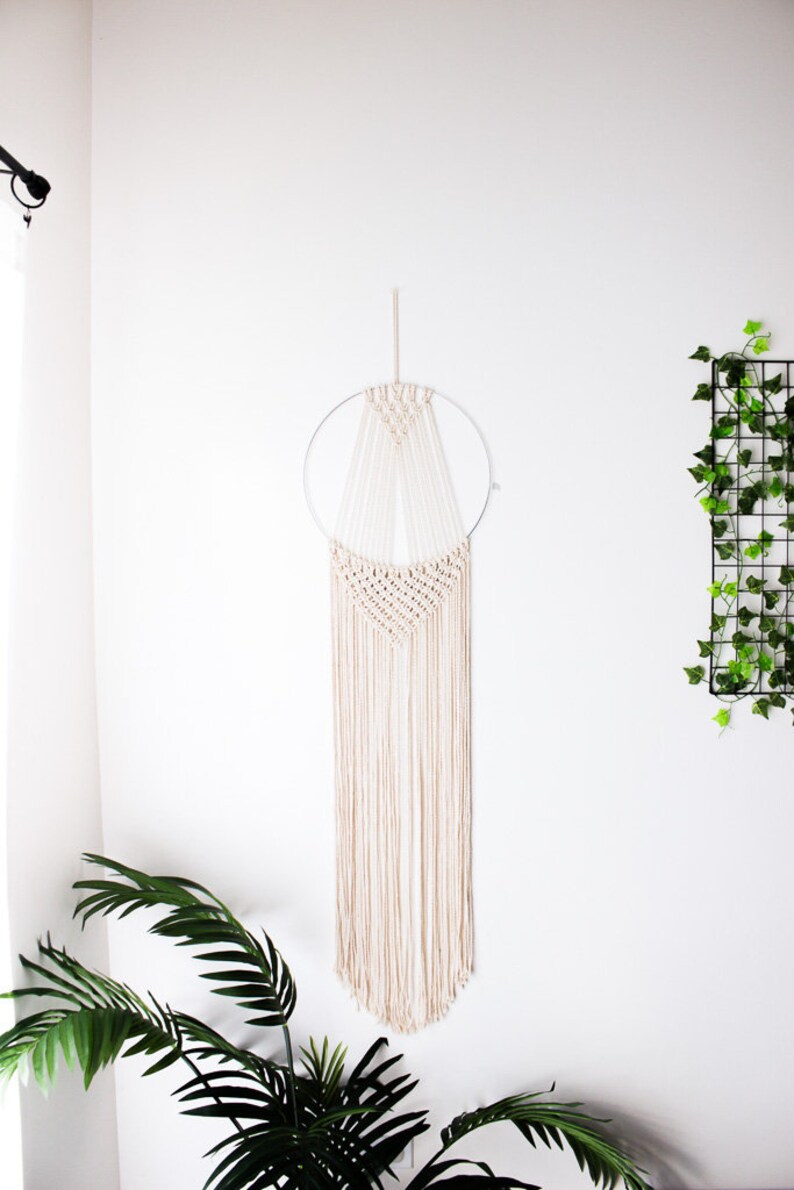 Op de afbeelding: Een cr&egrave;mekleurige macram&eacute; wandhanger met een cirkelvormig metalen frame. Het ontwerp heeft ingewikkelde knopen en lange, vloeiende franjes, waardoor een bohemien esthetiek ontstaat. De wanddecoratie wordt getoond tegen een witte muur, met een plant op de voorgrond.