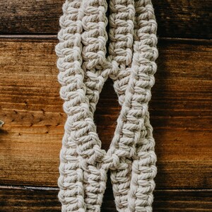 May include: A close-up of a macrame plant hanger made with natural cotton rope. The hanger is hanging against a wooden background.