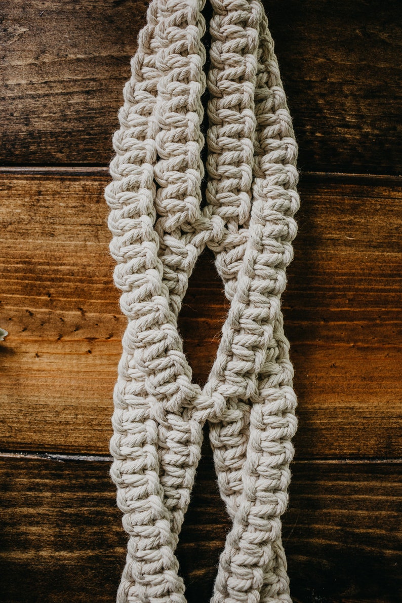 May include: A close-up of a macrame plant hanger made with natural cotton rope. The hanger is hanging against a wooden background.
