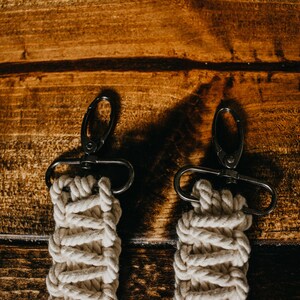 May include: Two white cotton rope keychains with black metal clasps. The keychains are hanging on a wooden surface.