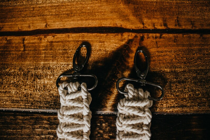 May include: Two white cotton rope keychains with black metal clasps. The keychains are hanging on a wooden surface.