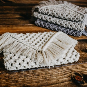 May include: Two crocheted coasters with fringe, one in a light beige color and the other in a darker gray color. The coasters are on a wooden surface.
