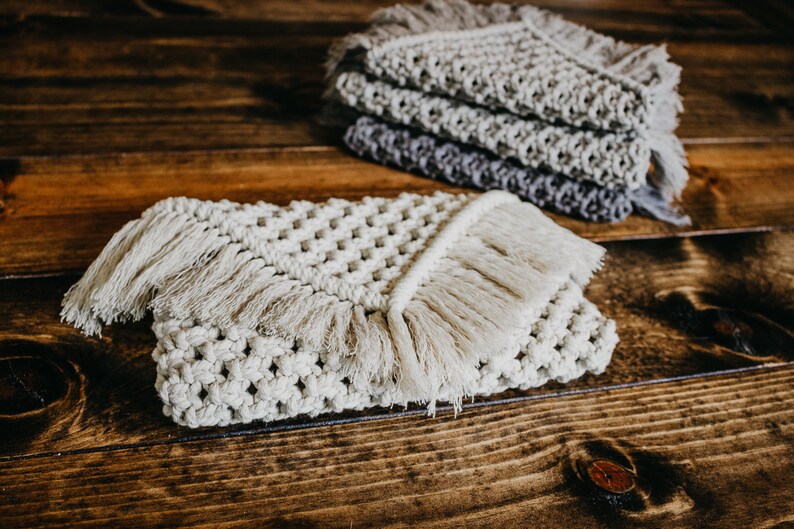 May include: Two crocheted coasters with fringe, one in a light beige color and the other in a darker gray color. The coasters are on a wooden surface.