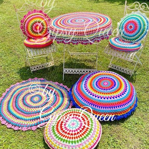 Op de afbeelding: Een set kleurrijke ronde kussens van gehaakt garen en een tafelkleed met een witte achtergrond en meerkleurige concentrische cirkels. De kussens zijn op een groene grasmat geplaatst.