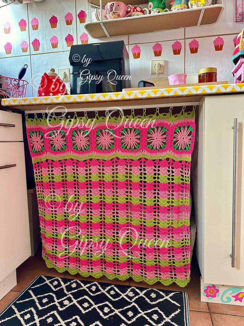 May include: A pink and green crocheted curtain with a scalloped bottom edge hangs from a white rod. The curtain is made of squares with flowers in the center. The text "by Gipsy Queen" is visible on the curtain.