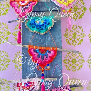 Op de afbeelding: Vijf gehaakte wandhangers in verschillende kleuren, waaronder roze, oranje, geel, blauw en groen, worden getoond op een houten achtergrond. Elke hanger heeft een cirkelvormig ontwerp met kralen en kwastjes.