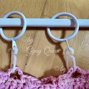 May include: Two white metal curtain rings with white plastic clips attached to a white metal curtain rod. The rings are hanging over a pink crocheted fabric.