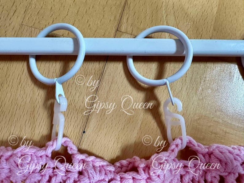 May include: Two white metal curtain rings with white plastic clips attached to a white metal curtain rod. The rings are hanging over a pink crocheted fabric.