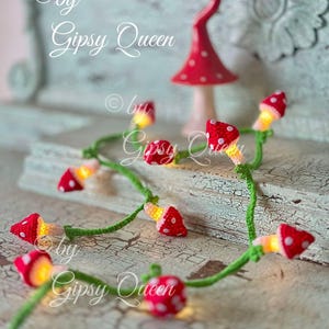 Puede incluir: Una cadena de luces con bombillas de ganchillo en forma de hongo. Los hongos son rojos con lunares blancos y tienen una luz amarilla en el interior. Las luces están enhebradas en un cable verde. El texto "by Gipsy Queen" es visible en la imagen.