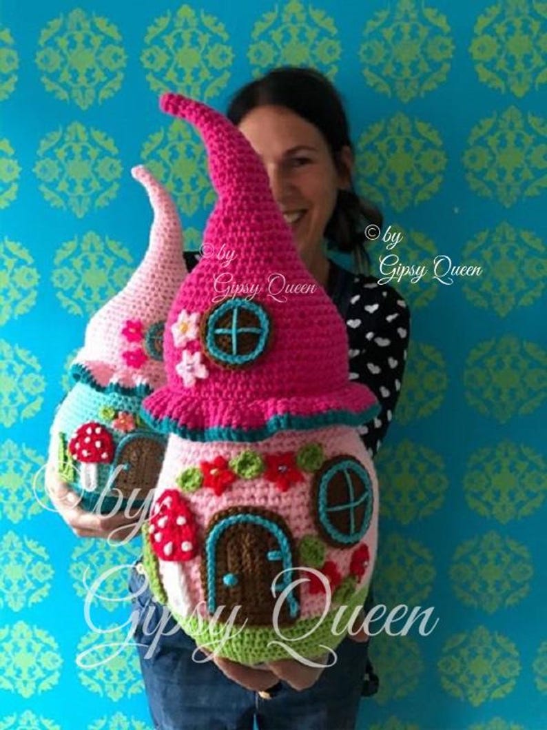 May include: Two crocheted fairy houses, one pink and one light pink, with brown doors, blue windows, and mushroom accents. The houses are held by a person, and the text "by Gipsy Queen" is visible.