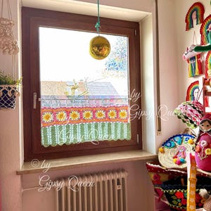 May include: A window with a crocheted valance featuring a floral design in shades of pink, yellow, and green. The valance is hanging above a white radiator. A disco ball hangs from the ceiling above the window.