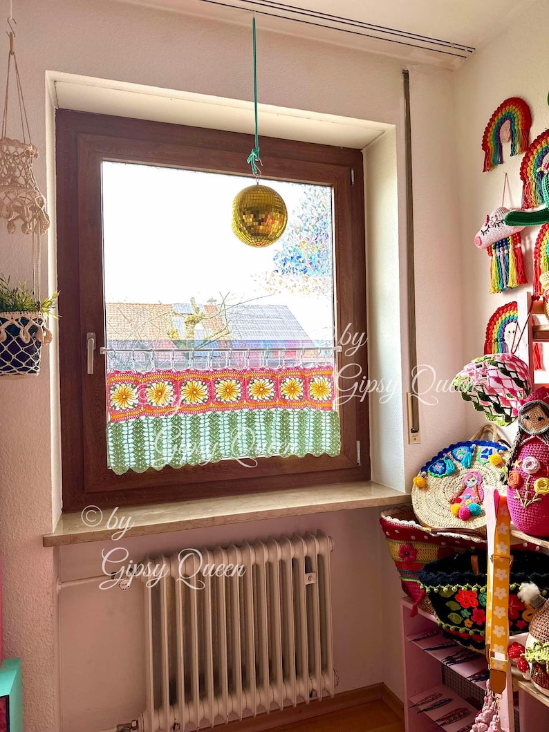 May include: A window with a crocheted valance featuring a floral design in shades of pink, yellow, and green. The valance is hanging above a white radiator. A disco ball hangs from the ceiling above the window.