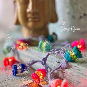 May include: A string of colourful crocheted flower lights with warm white LED bulbs. The lights are strung on a purple cord and are lit up. The lights are on a wooden surface with a blurred image of a golden Buddha statue in the background. The text "© by Gipsy Queen" is visible in the image.