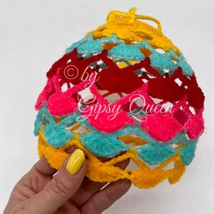 May include: A crocheted ball ornament with a yellow, turquoise, pink, and red color scheme. The ornament is made of yarn and has a textured surface. The text "by Gipsy Queen" is visible on the ornament.