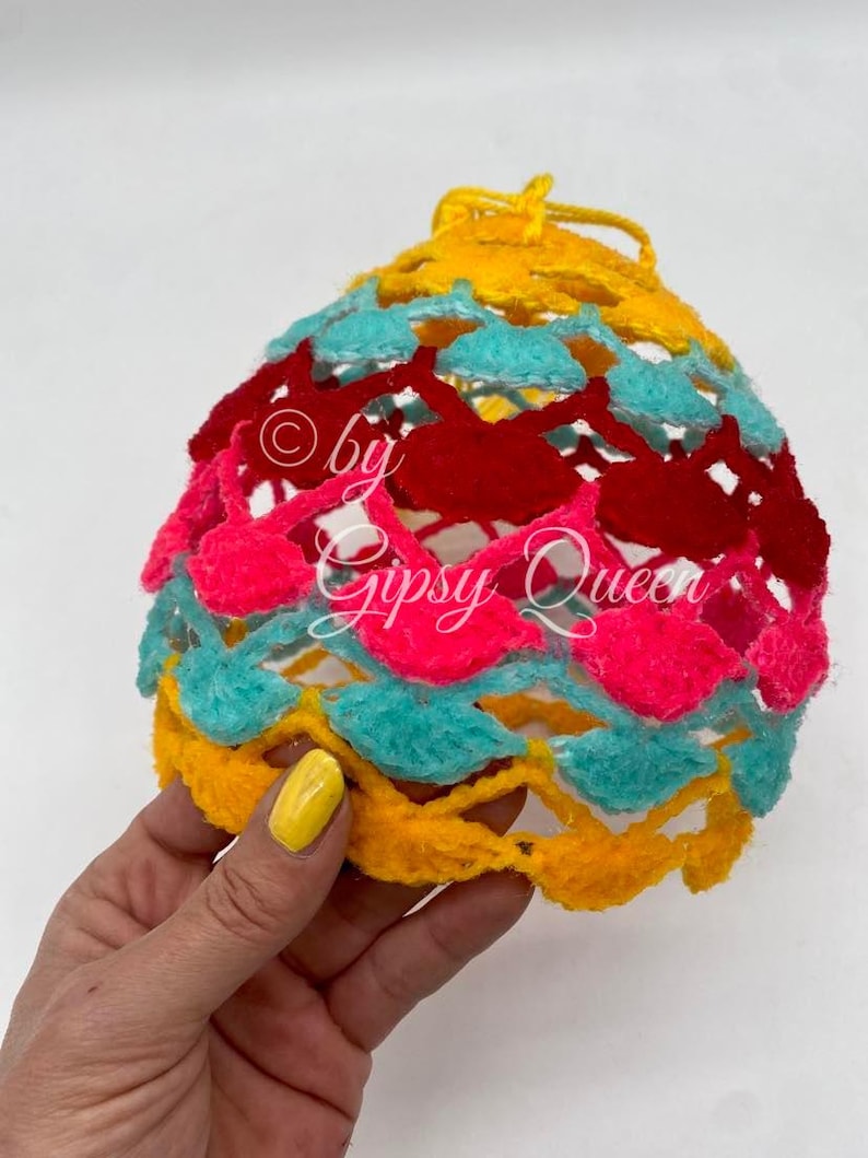 May include: A crocheted ball ornament with a yellow, turquoise, pink, and red color scheme. The ornament is made of yarn and has a textured surface. The text "by Gipsy Queen" is visible on the ornament.