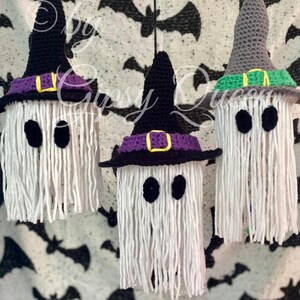 K&ouml;nnte beinhalten: Drei handgefertigte Halloween-Geisterdekorationen. Jeder Geist besteht aus wei&szlig;em Garn mit schwarzen ovalen Augen und einem geh&auml;kelten Hexenhut. Die H&uuml;te sind schwarz, grau und lila mit gelben und gr&uuml;nen Akzenten. Der Hintergrund ist ein schwarz-wei&szlig;es Fledermausmuster.