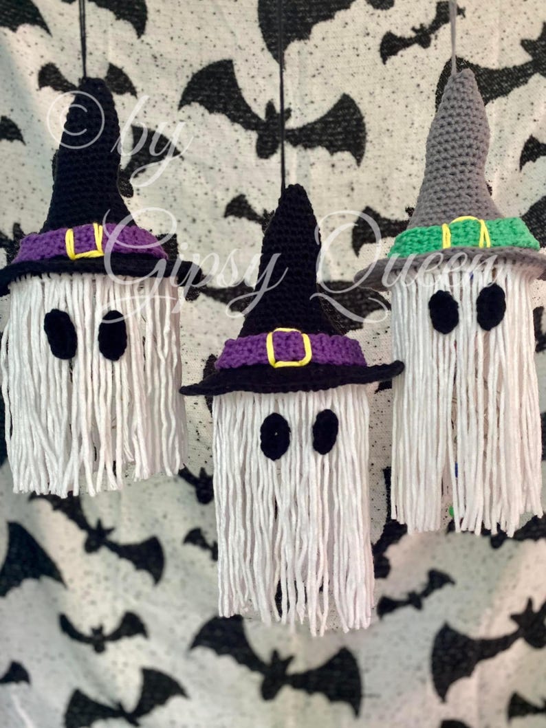 K&ouml;nnte beinhalten: Drei handgefertigte Halloween-Geisterdekorationen. Jeder Geist besteht aus wei&szlig;em Garn mit schwarzen ovalen Augen und einem geh&auml;kelten Hexenhut. Die H&uuml;te sind schwarz, grau und lila mit gelben und gr&uuml;nen Akzenten. Der Hintergrund ist ein schwarz-wei&szlig;es Fledermausmuster.