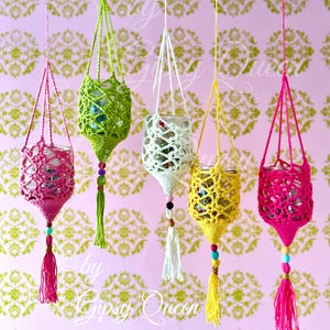 Puede incluir: Cinco coloridos portavasos de ganchillo colgando sobre un fondo rosa con un estampado floral. Cada soporte es de un color diferente: rosa, verde, blanco, amarillo y rosa. Cada uno tiene borlas y cuentas, y el texto "by Gipsy Queen".