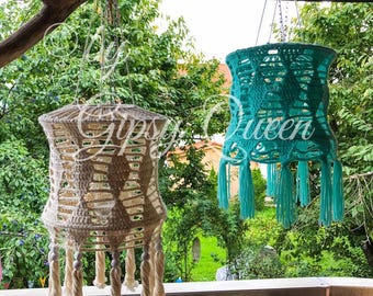 May include: Two crocheted macrame hanging light fixtures, one in beige and one in teal, with tassels hanging from the bottom. The lights are hanging from a wooden branch and are in front of a green leafy background.