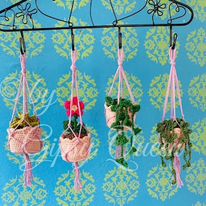 以下が含まれることがあります： ピンクの糸と緑の植物を使った、4つの編まれた吊り下げ式プランター。プランターは、ハート型の切り抜きのある黒い金属製のラックに吊り下げられています。
