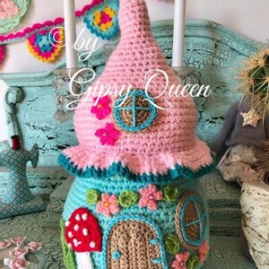 May include: A crocheted fairy house with a pink roof, teal walls, a brown door, and a mushroom with red and white spots. The house is decorated with flowers and a window. The text "by Gipsy Queen" is visible in the image.