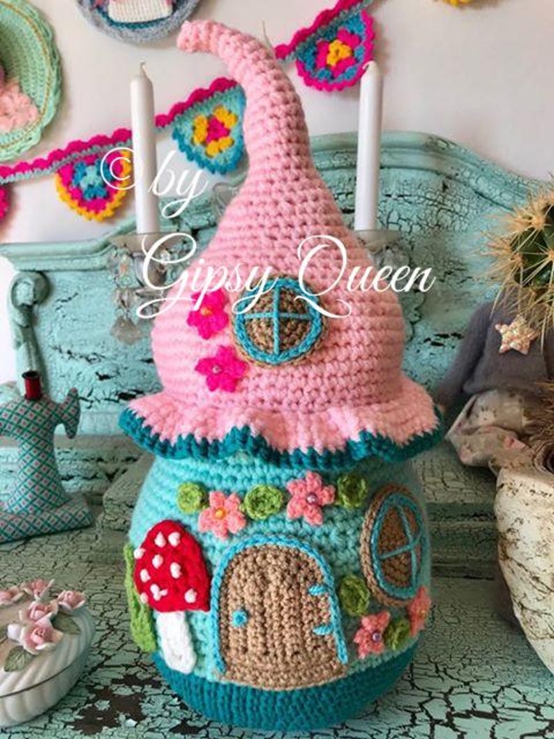 May include: A crocheted fairy house with a pink roof, teal walls, a brown door, and a mushroom with red and white spots. The house is decorated with flowers and a window. The text "by Gipsy Queen" is visible in the image.