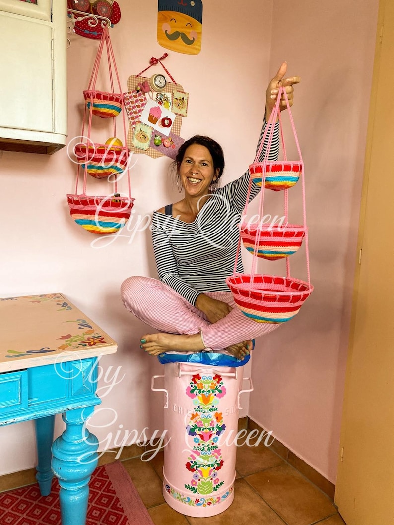 May include: Three hanging baskets made of crocheted yarn in pink, red, and yellow stripes. The baskets are hanging from a pink wall and are being held by a person. The baskets are empty.