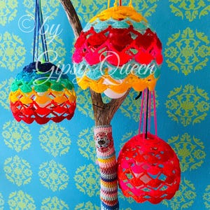 May include: Three colorful crocheted lampshades hanging from a branch wrapped in colorful yarn. The lampshades are made of a rainbow of colors, including red, orange, yellow, green, blue, and purple.