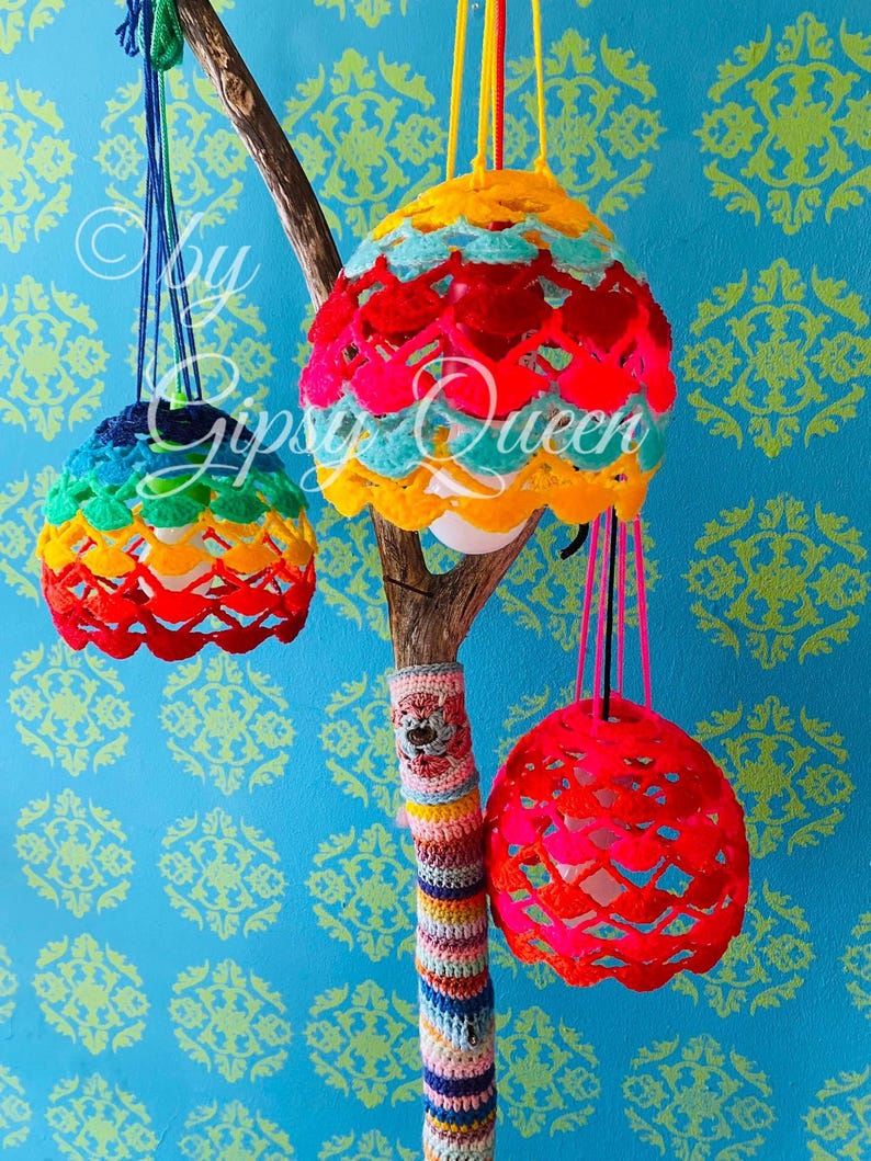 May include: Three colorful crocheted lampshades hanging from a branch wrapped in colorful yarn. The lampshades are made of a rainbow of colors, including red, orange, yellow, green, blue, and purple.