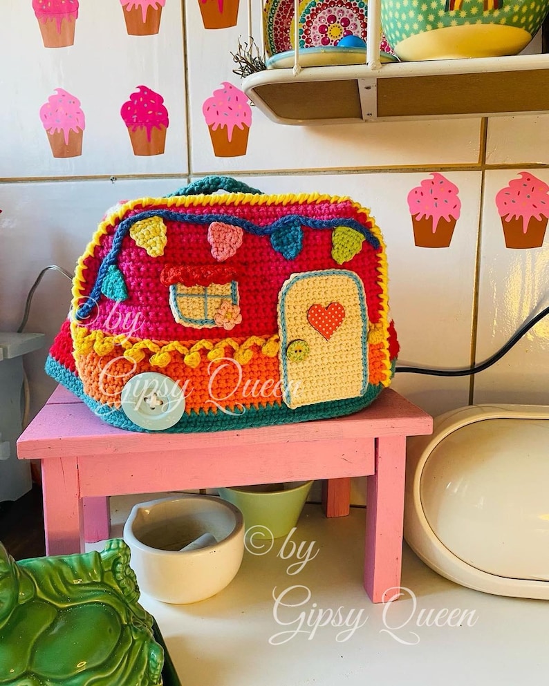 May include: A colorful, crocheted camper with a door featuring a heart design, sitting on a pink table. The camper is decorated with various colors and shapes. The text "by Gipsy Queen" is visible.