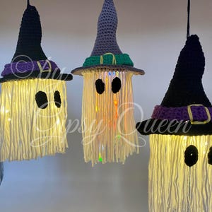 K&ouml;nnte beinhalten: Drei h&auml;ngende Halloween-Dekorationen in Form von Geistern. Jeder Geist hat einen geh&auml;kelten Hexenhut in Schwarz, Grau oder Lila. Die Geister bestehen aus cremefarbenem Garn mit schwarzen Augenausschnitten und sind mit Lichtern beleuchtet.