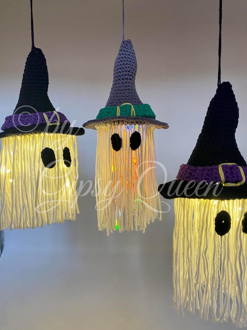 K&ouml;nnte beinhalten: Drei h&auml;ngende Halloween-Dekorationen in Form von Geistern. Jeder Geist hat einen geh&auml;kelten Hexenhut in Schwarz, Grau oder Lila. Die Geister bestehen aus cremefarbenem Garn mit schwarzen Augenausschnitten und sind mit Lichtern beleuchtet.