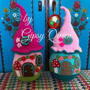 May include: Two crocheted gnome houses, one pink and one blue, with a whimsical design featuring flowers, mushrooms, and windows. The houses are standing in front of a blue door with floral designs.